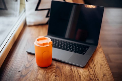 A laptop opened and placed next to an orange thermos cup on a wooden table, indicative of remote