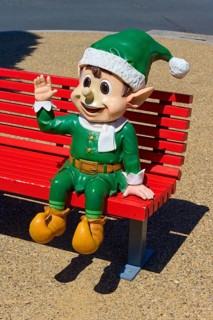 Elf figure in green costume greeting on a red bench in sunny weather, Bethlehem, Tauranga, Bay of