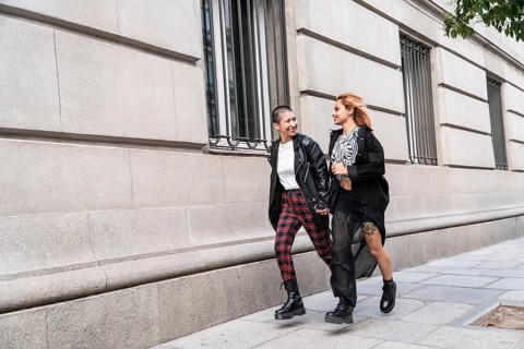 A trendy lesbian couple walks hand in hand along a city street. Both are dressed stylishly in