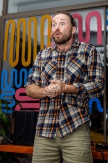 A young Caucasian man with a beard and long hair stands with his arms outstretched. He wears a