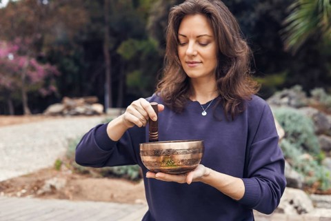 A young Caucasian woman with brown hair plays a singing bowl outdoors. She wears a dark sweater and