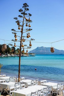Tables lined up on the beach with turquoise water and boats in the background on a sunny day