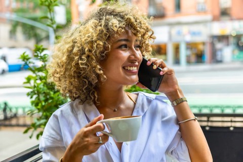 Smiling woman with curly hair talking on a mobile phone and drinking coffee at an outdoor cafe,
