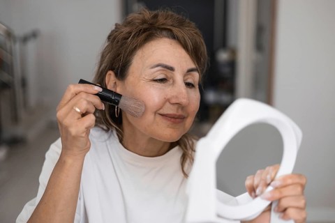 A middle aged woman sits comfortably in bright room at home, using brush to apply makeup on her