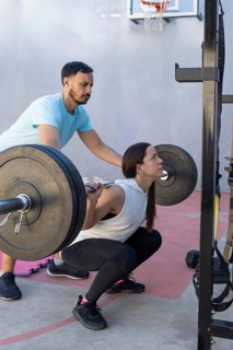 Personal trainer providing guidance to a woman executing barbell squats in an outdoor gym,
