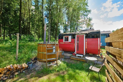 Small red sauna and hot tub in green forest surrounded by trees. Stack of firewood and chairs for