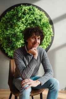 A young man with curly hair sits thoughtfully, framed by a lush green wall. This image emphasizes