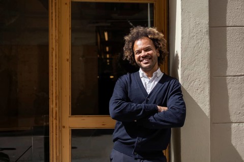 A cheerful man with curly hair smiles confidently while standing outside a modern coworking space