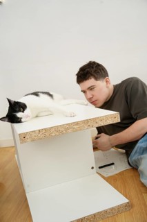 A young man assembles a white flatpack furniture piece, while a black and white cat reclines beside