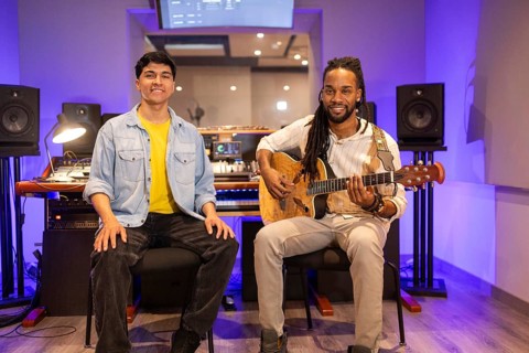 Two musicians in a modern recording studio. One playing an acoustic guitar, the other seated. The