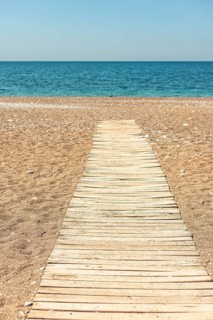 A wooden walkway leads to a calm beach with golden sand and clear blue water. The sky is bright and