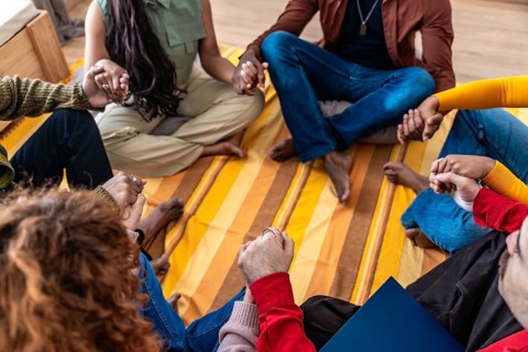 Diverse group sitting on a blanket in a circle, holding hands and sharing support during a therapy