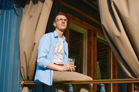 Middle-aged man sits on balcony in casual clothes holding drink. He looks out towards green trees