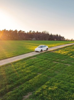 A white car on a rural road at sunset, surrounded by green meadows and a forest in the background,
