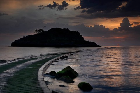 Silhouette of a small rocky island in the Atlantic Ocean, onto which a half-flooded causeway leads,