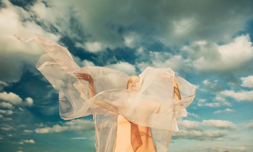 A woman is wrapped in sheer, flowing fabric against a vast, cloudy sky. The fabric catches the