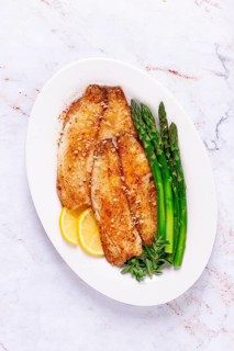 Fresh fish filets are served on a white plate alongside green asparagus and slices of lemon. The