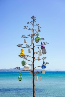 A coastal art installation features a tree adorned with colorful decorations, set against a bright