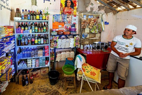 A small grocery store with shelves full of goods. A man wearing a cap stands relaxed next to it.