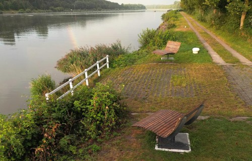 A quiet river with green riparian vegetation and several wooden benches to relax on, ergonomic