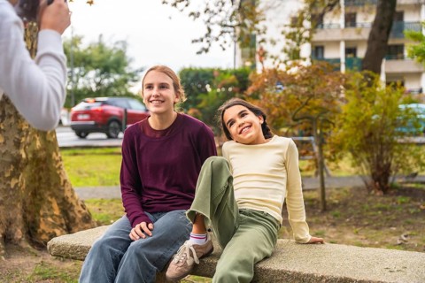 Two sisters smiling and relaxing on a park bench while a family member photographs their candid,