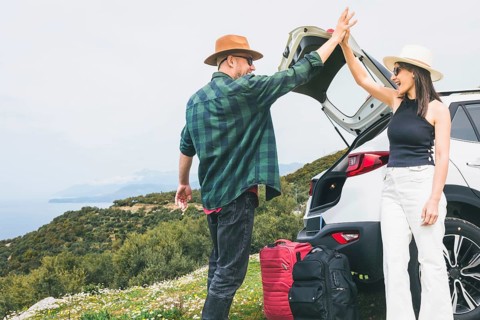 Happy Travelers tourist couple hi5 stand put luggage to white rented SUV vehicle trunk on scenic