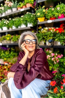 Mature woman with gray hair and glasses smiling thoughtfully, surrounded by colorful potted