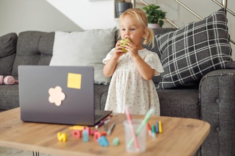 A young Caucasian girl with blonde hair stands by a laptop on a wooden table. Colorful toys and