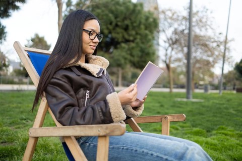 A Hispanic woman with glasses enjoys reading a book while sitting comfortably in a park. She