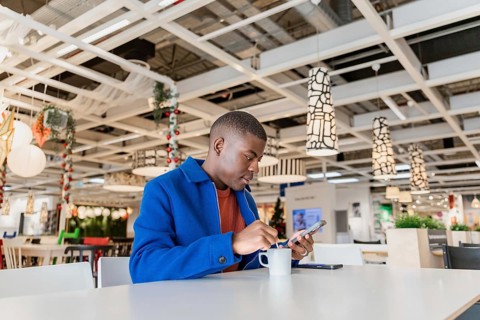 A young Black man using a phone and holding a cup and sits at a table in a modern cafe, The
