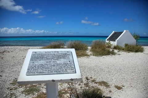 Information board on the pebble beach with a small white house and turquoise sea in the background