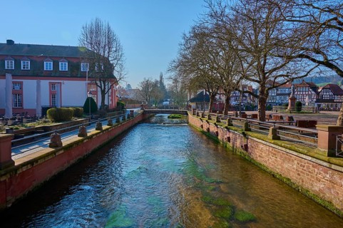 Quiet river flows between trees and historic buildings under blue spring skies, Mümling river,