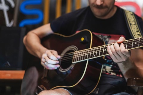 A young Caucasian man with a beard plays an acoustic guitar. He wears a black t-shirt and is