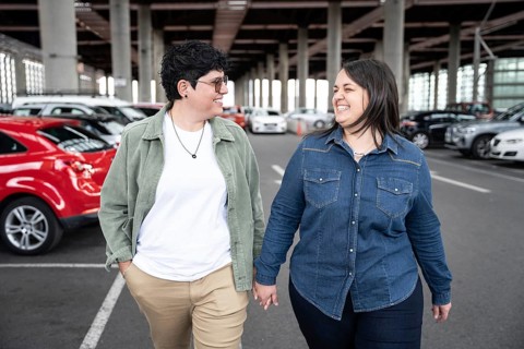 Lesbian couple laughing and walking hand in hand through a parking area. They are dressed casually,