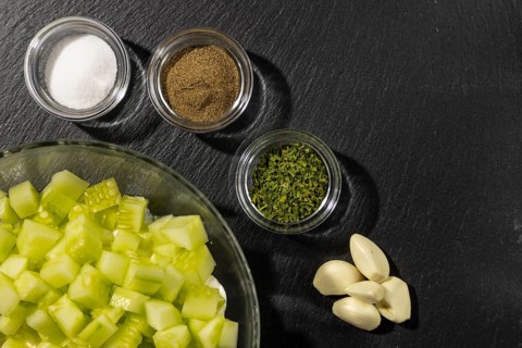 Bowl with sliced cucumber and sour cream, spices and garlic next to it