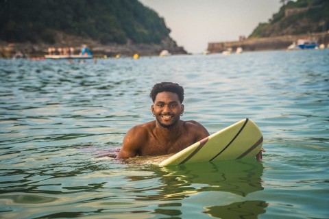 Young black man in the ocean smiling at the camera, holding a surfboard on a sunny summer day,