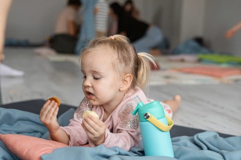 A young Caucasian girl with light brown hair enjoys snacks while sitting on a mat. The background
