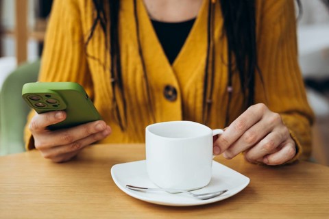 A woman in a cozy yellow sweater sits at a cafe table, holding a white coffee cup in one hand and a