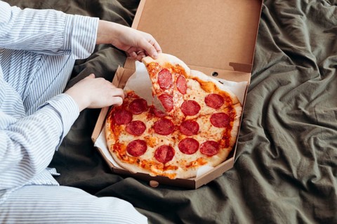 Cropped unrecognizable woman indulges in a cheesy pepperoni pizza while lying in bed, depicted in a