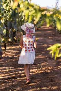A young girl, wearing a sun hat and a white dress with red and orange accents, walks through a