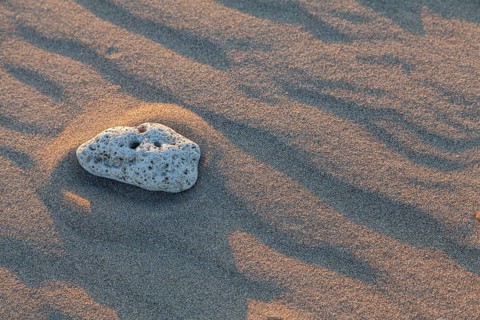 A single stone on the sandy beach, surrounded by soft shadows and wave patterns in the light of the