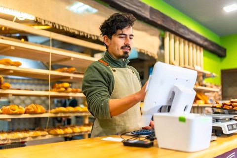 Latin male young salesman wearing apron using touchscreen computer in the counter of bakery shop