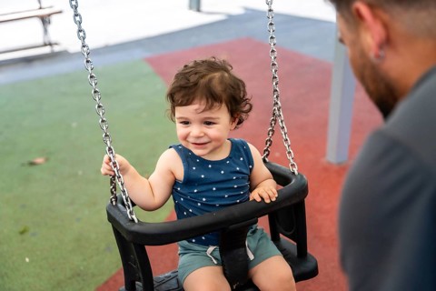 Happy infant child sitting in a safety swing, looking at parent and smiling widely during playtime,