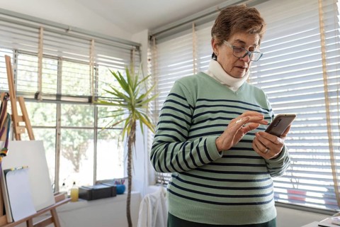 Older woman wearing blue reading glasses checks her smartphone in a bright home studio, with a