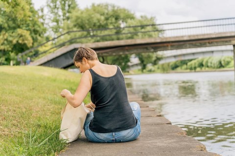 Back view of woman looking into tote bag while sitting on urban riverbank, A woman sits casually by