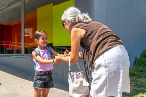 Grandmother is greeting her cheerful granddaughter, wearing a backpack, outside a modern school
