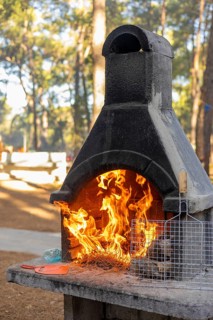 A stone outdoor fireplace with flames rising from the chimney. The setting is surrounded by trees,