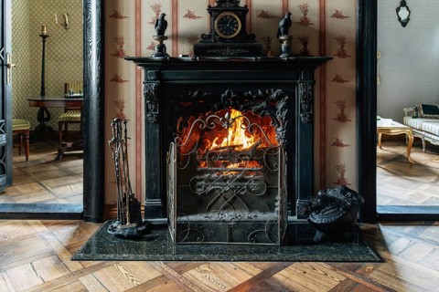 A vintage room features an ornate dark fireplace with a bright burning fire and a wrought iron
