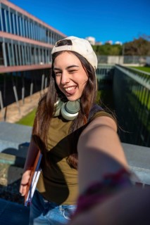 Young female student smiling and winking for a selfie on campus, holding books with headphones