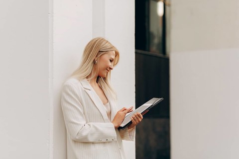A businesswoman in a white blazer stands outside against a wall, looking at a digital tablet. She